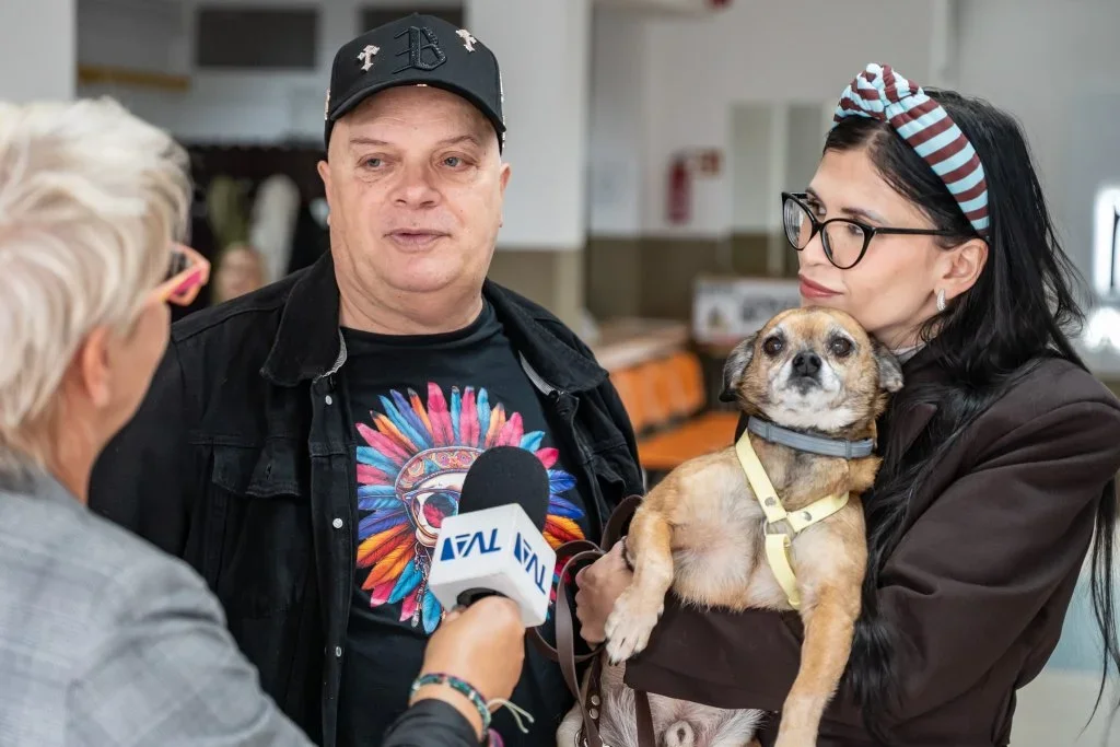 Mężczyzna i kobieta trzymająca małego psa udzielają wywiadu dziennikarce z mikrofonem oznaczonym logo TVN w pomieszczeniu o charakterze publicznym.