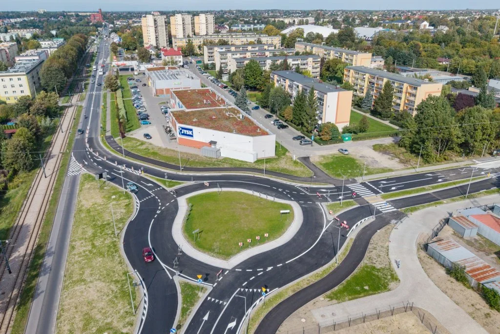 Widok z lotu ptaka na nowo wybudowane rondo miejskie w dzielnicy z blokami mieszkalnymi, sklepem JYSK i torowiskiem tramwajowym.