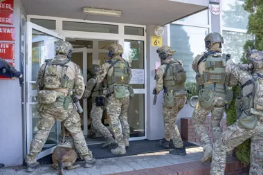 Uzbrojeni funkcjonariusze polskiej policji w kamuflażu wchodzą do budynku przy ul. Szkolnej 6A, jeden z nich prowadzi psa służbowego.