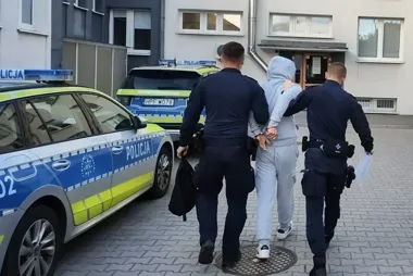 Dwóch policjantów prowadzi w kajdankach mężczyznę w szarym dresie z kapturem w stronę policyjnych radiowozów przed budynkiem mieszkalnym.