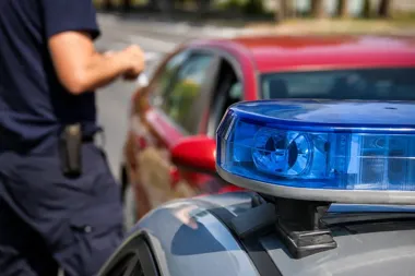 Mundurowy funkcjonariusz stoi przy policyjnym radiowozie z niebieskim światłem, w tle czerwone auto na drodze.