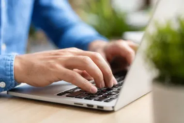 Mężczyzna w niebieskiej koszuli pisze na klawiaturze laptopa stojącego na drewnianym blacie.